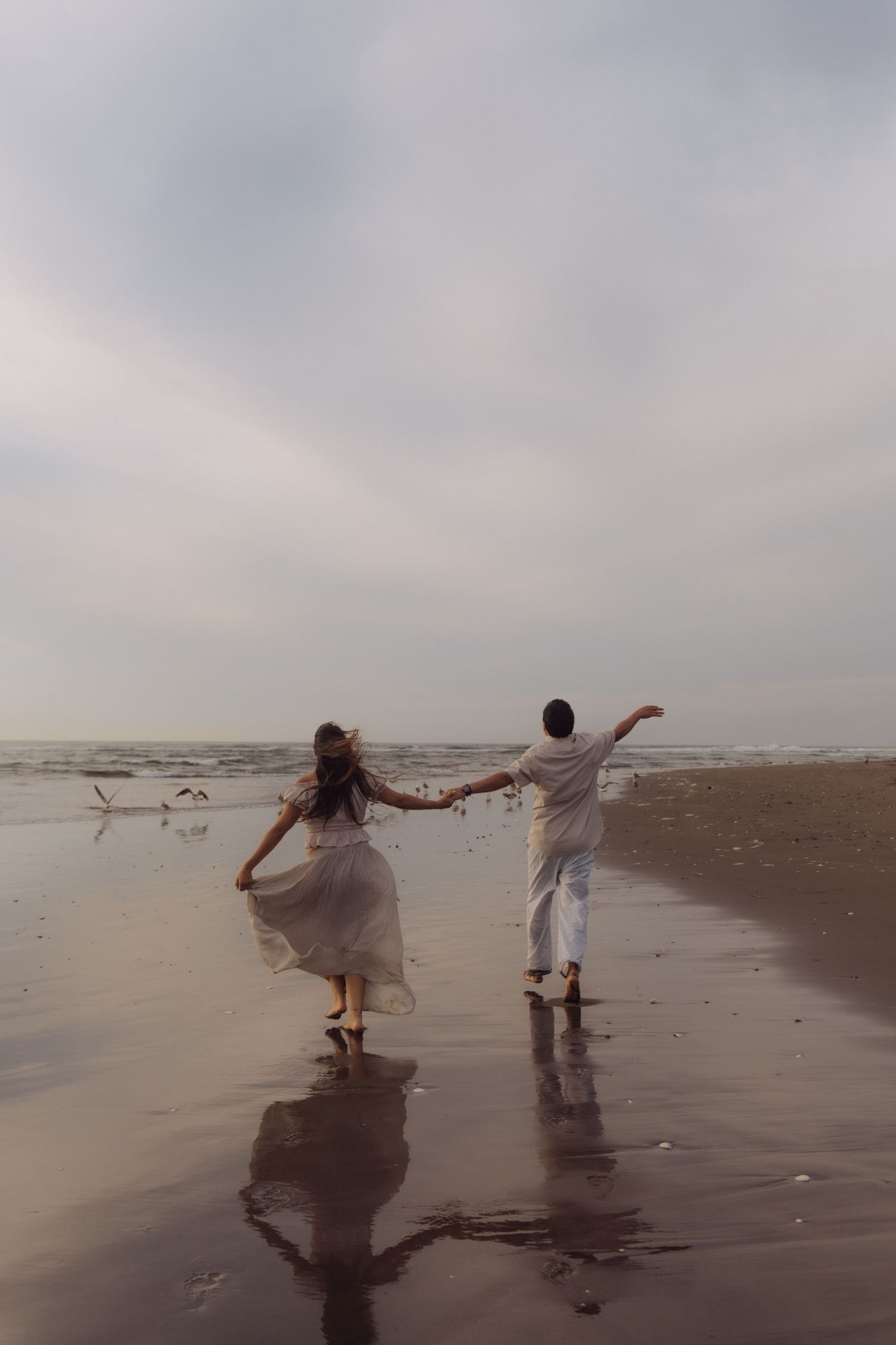Couple holding hands walking on reflective wet sand beach with seagulls and ocean waves.