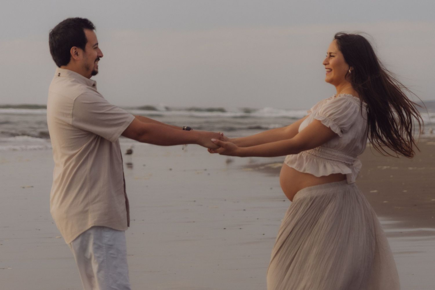 Smiling pregnant couple holding hands and dancing together on a beach at golden hour.