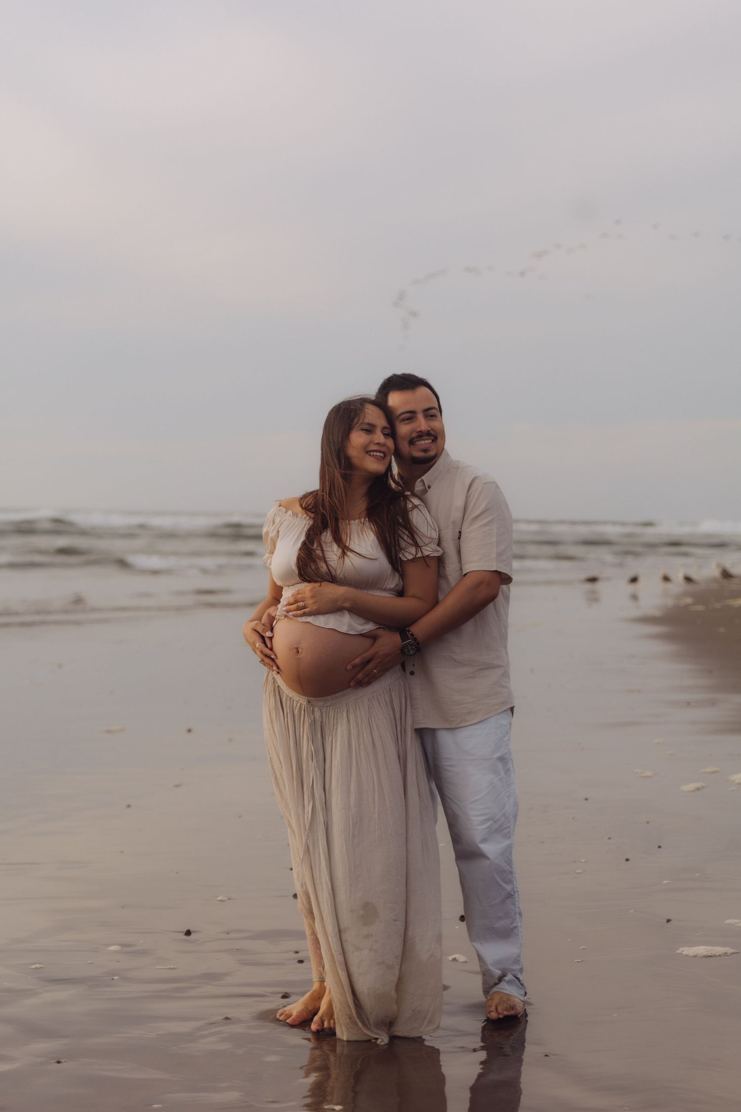 Happy couple embracing on beach, man holding pregnant woman in flowing beige maternity dress.