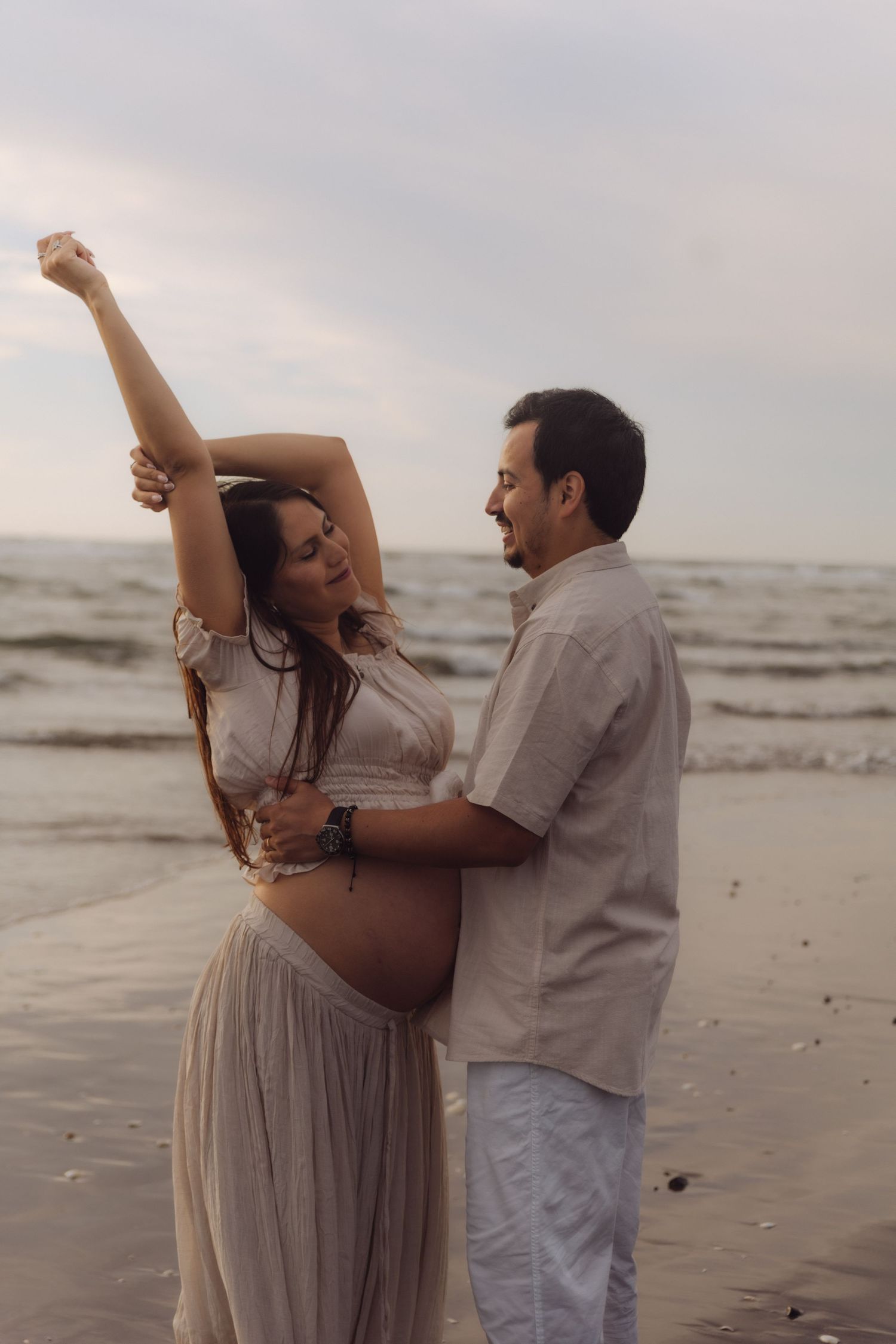 Pregnant couple dancing on beach at sunset, woman raising arm gracefully near ocean waves.