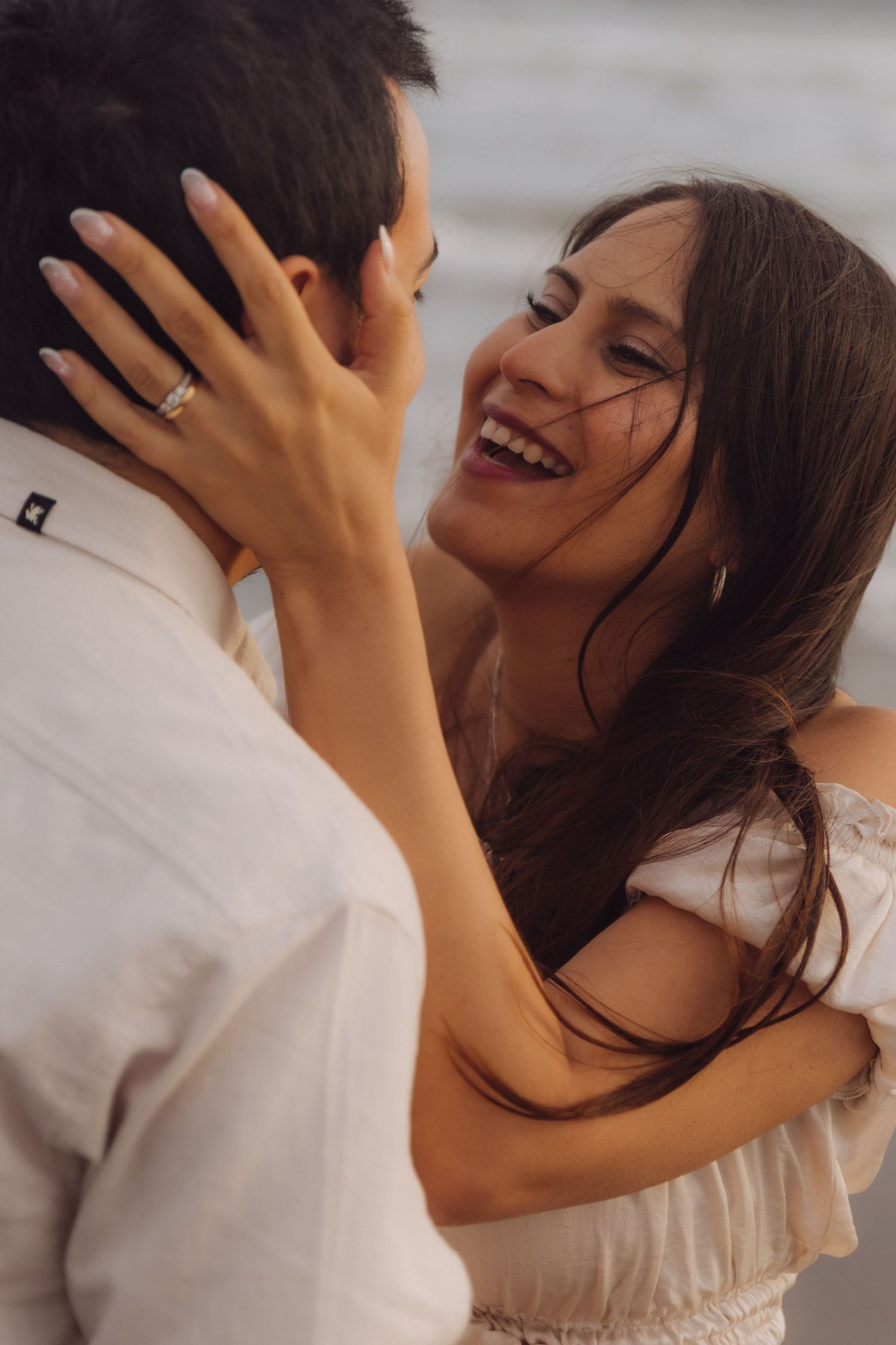 Smiling woman with dark hair embraces man on beach, wearing white off-shoulder dress and engagement ring.