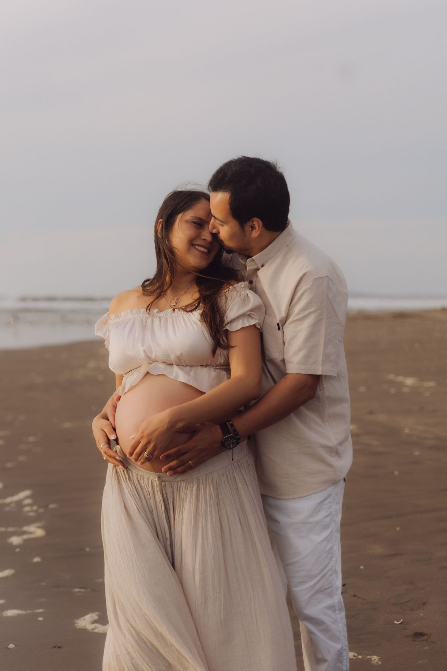 Pregnant couple embracing on beach at sunset, man kissing woman's head while holding her baby bump.