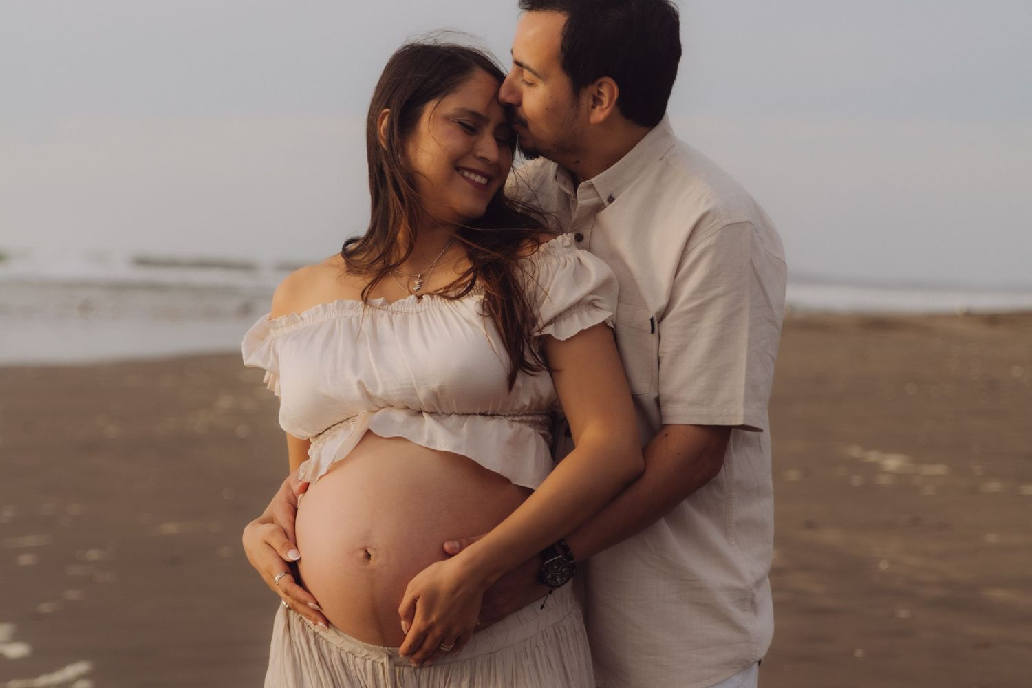 Happy pregnant couple cuddling on sandy beach, both cradling the baby bump with ocean in background.