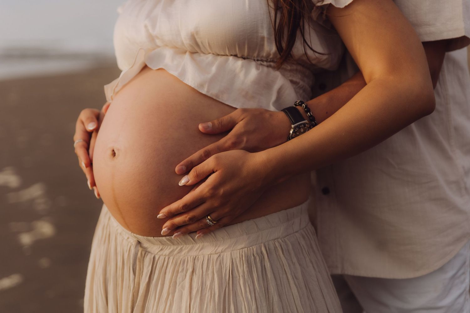 Close-up of pregnant woman's bare baby bump being tenderly held by couple on beach.