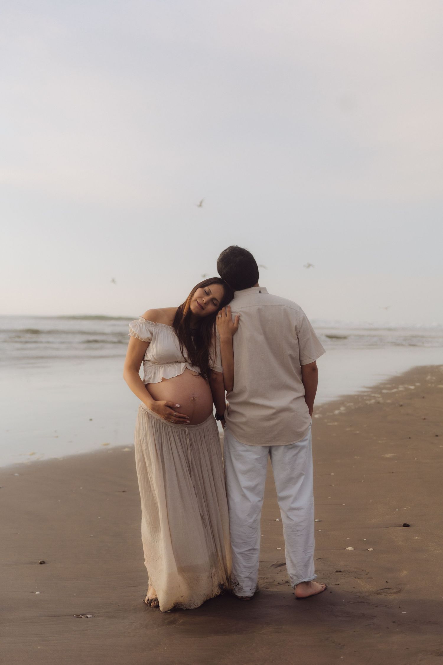 Pregnant woman in flowy dress leans on partner's shoulder while holding belly on beach at dusk.
