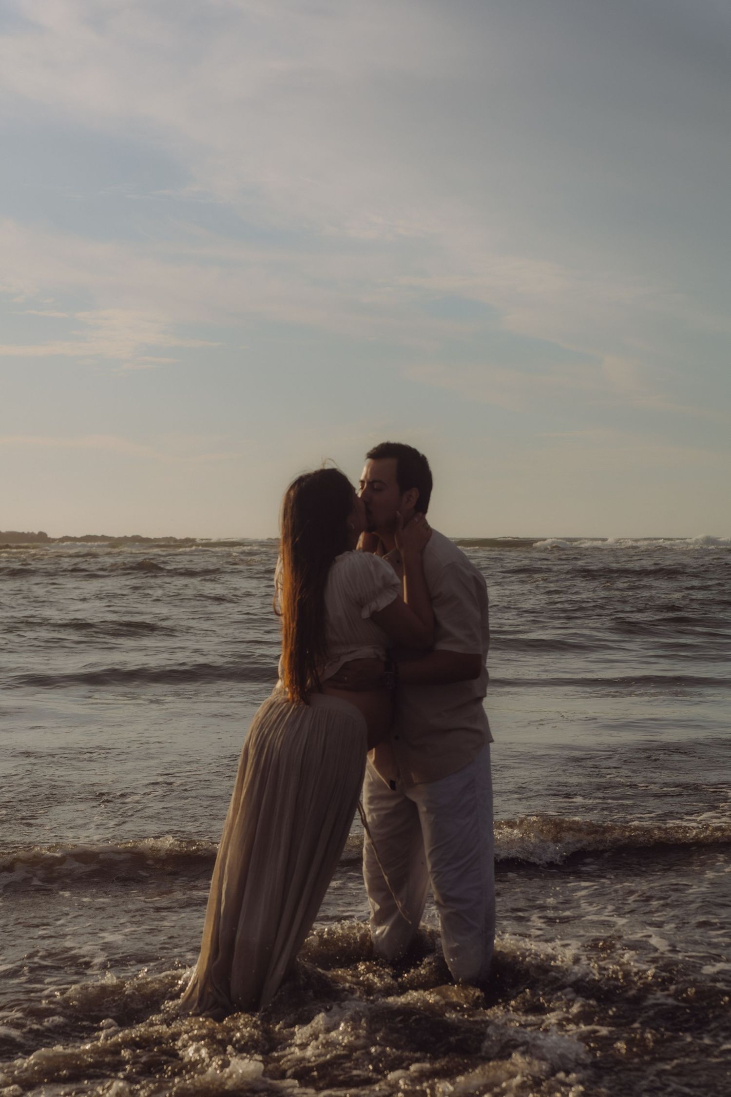 Couple sharing a romantic kiss while standing in ocean waves at sunset on the beach.