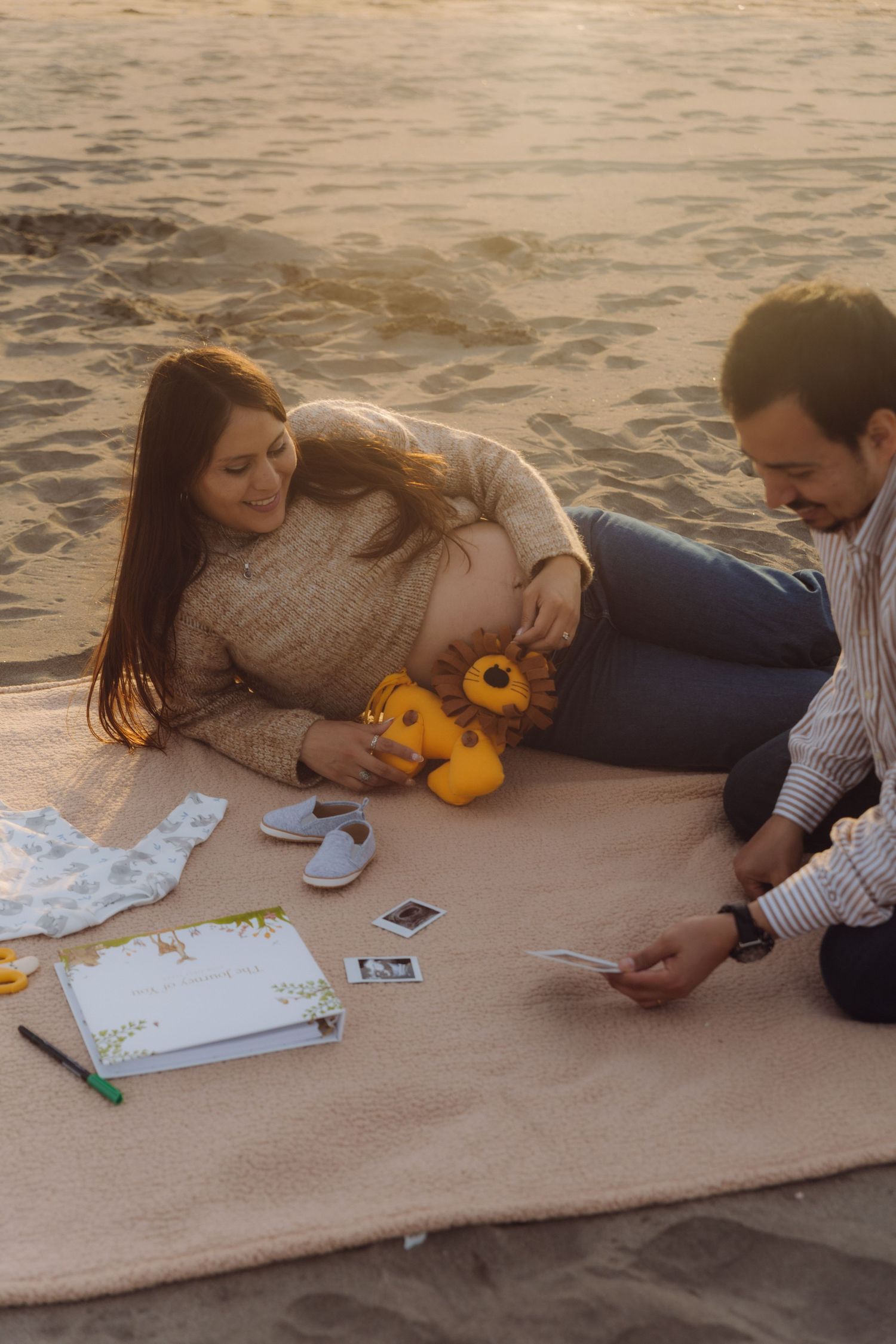 Pregnant couple sitting on beach blanket reviewing ultrasound photos with baby toys and book.