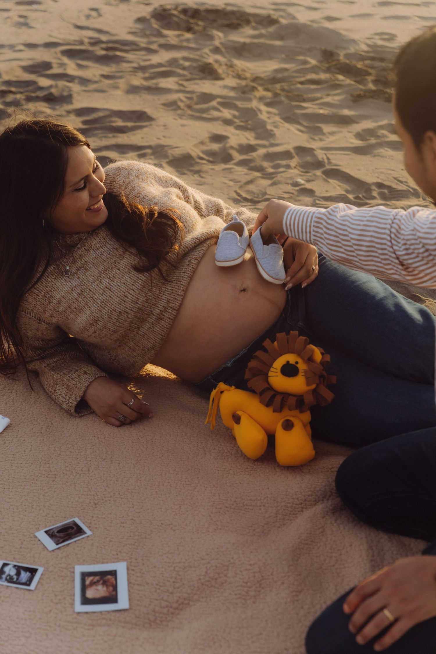 Pregnant woman lying on beach blanket as partner holds tiny baby shoes over her bare bump.