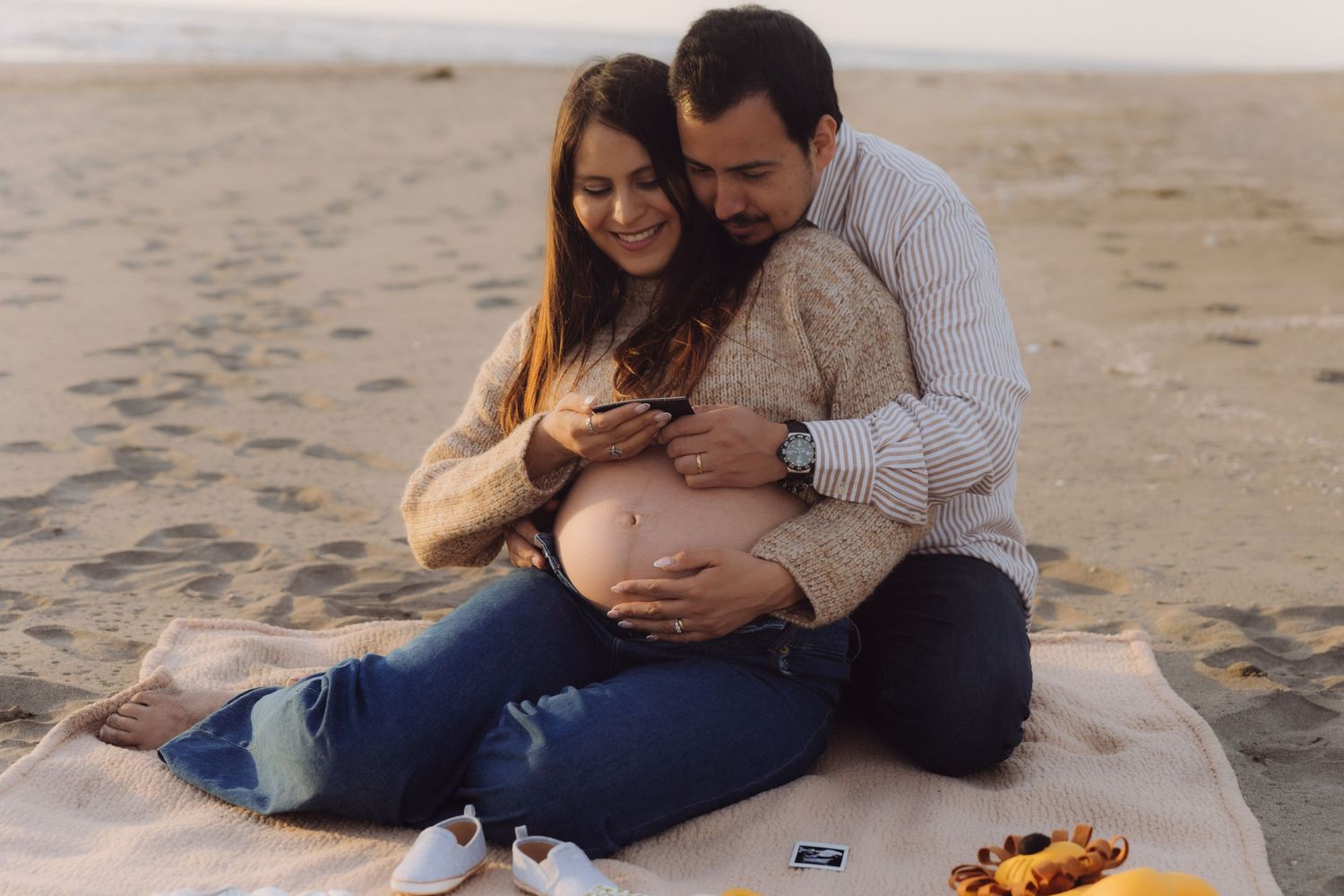 Couple sitting on beach blanket, man tenderly embracing pregnant woman from behind at sunset.