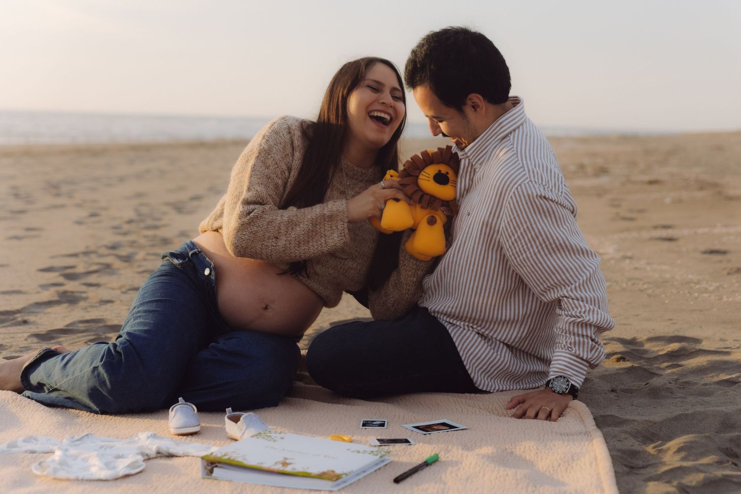 Laughing pregnant couple sitting on beach blanket playing with stuffed lion baby toy together.