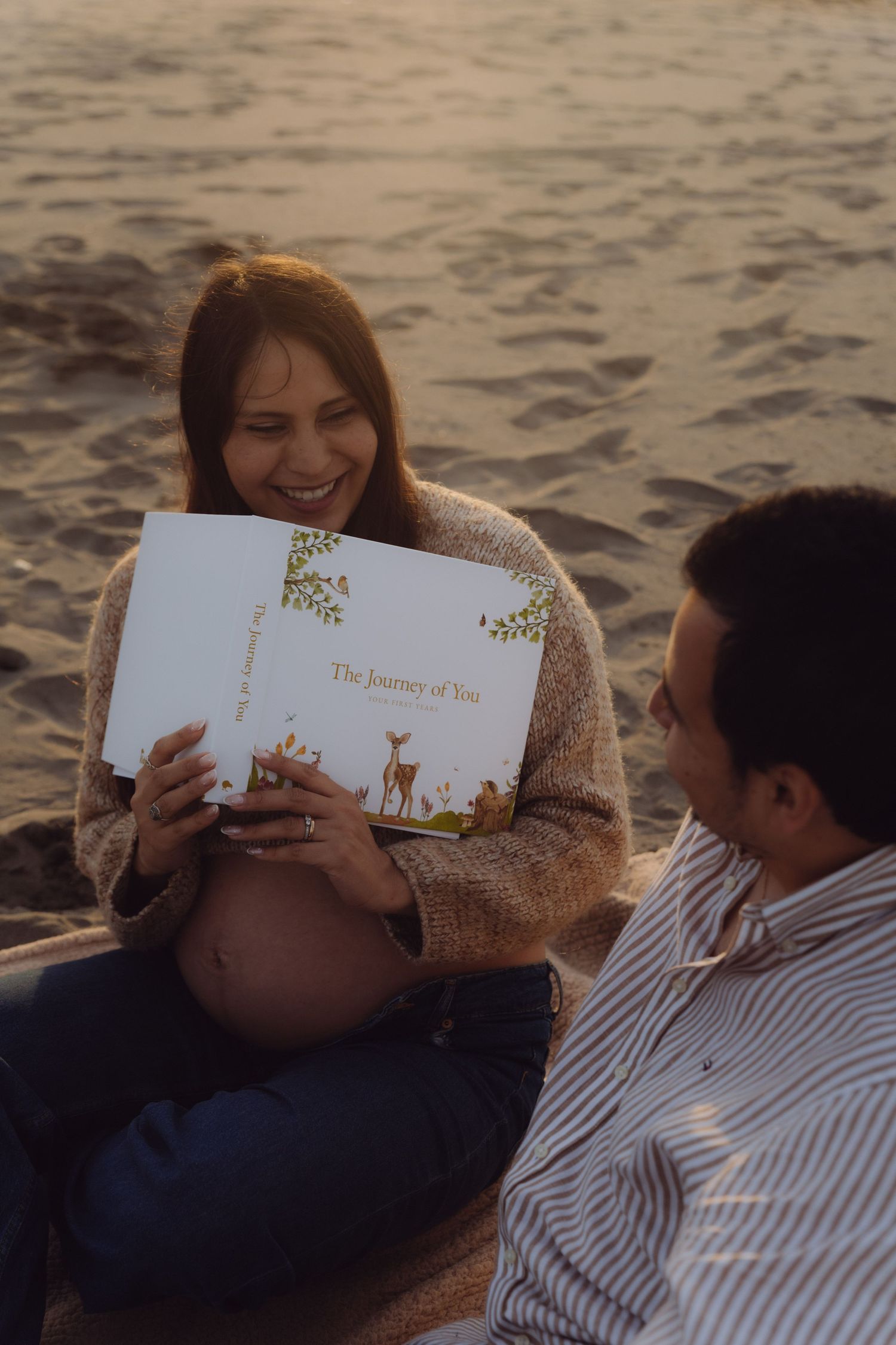 Pregnant woman sitting on beach sand reading a baby book while smiling, with her partner beside her at sunset.