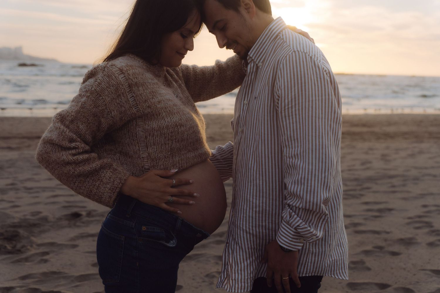 Expecting couple standing on beach at sunset, tenderly touching foreheads while cradling pregnant belly.