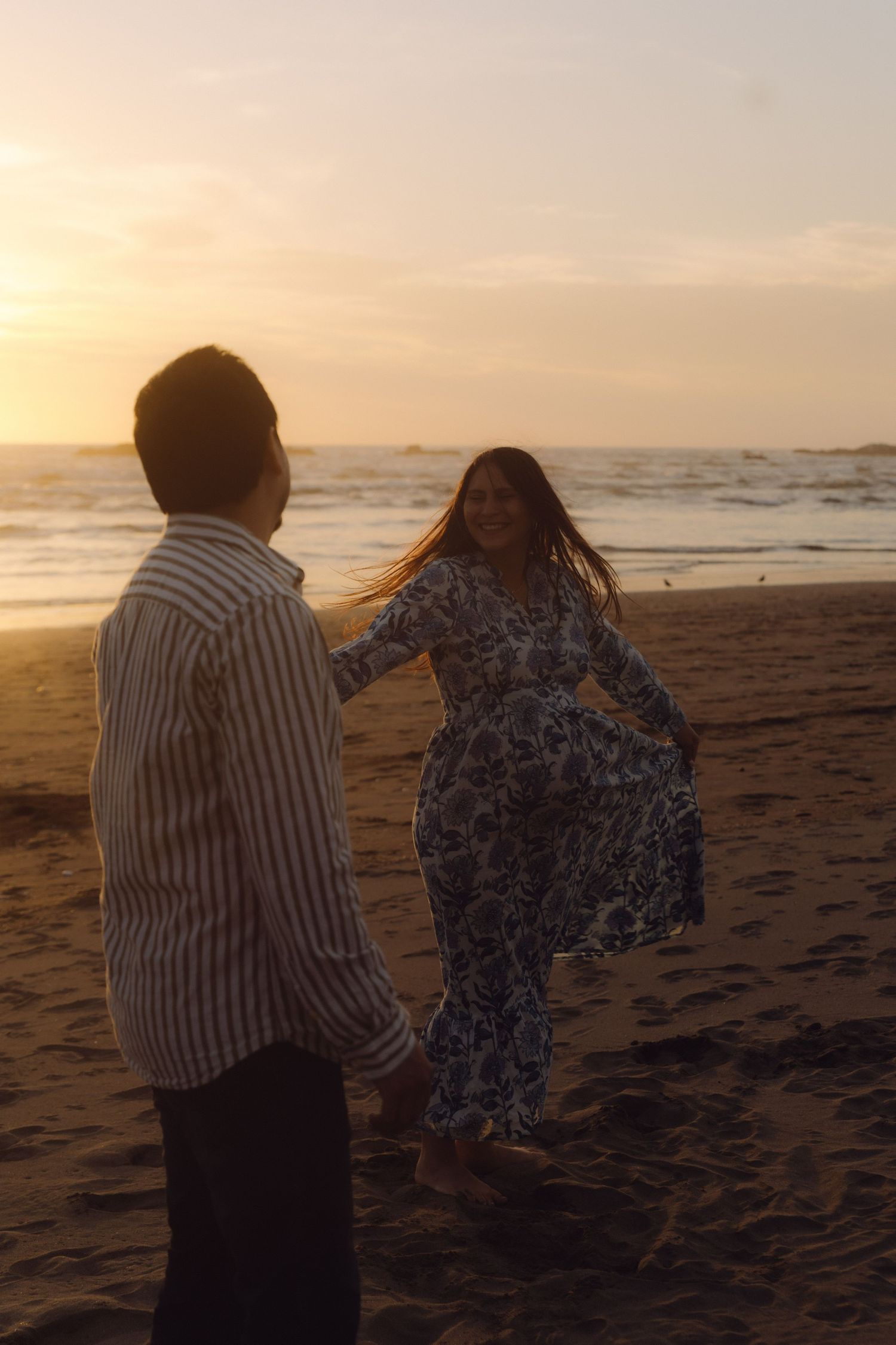 Pregnant woman in floral dress dancing joyfully with partner on sandy beach during golden sunset.