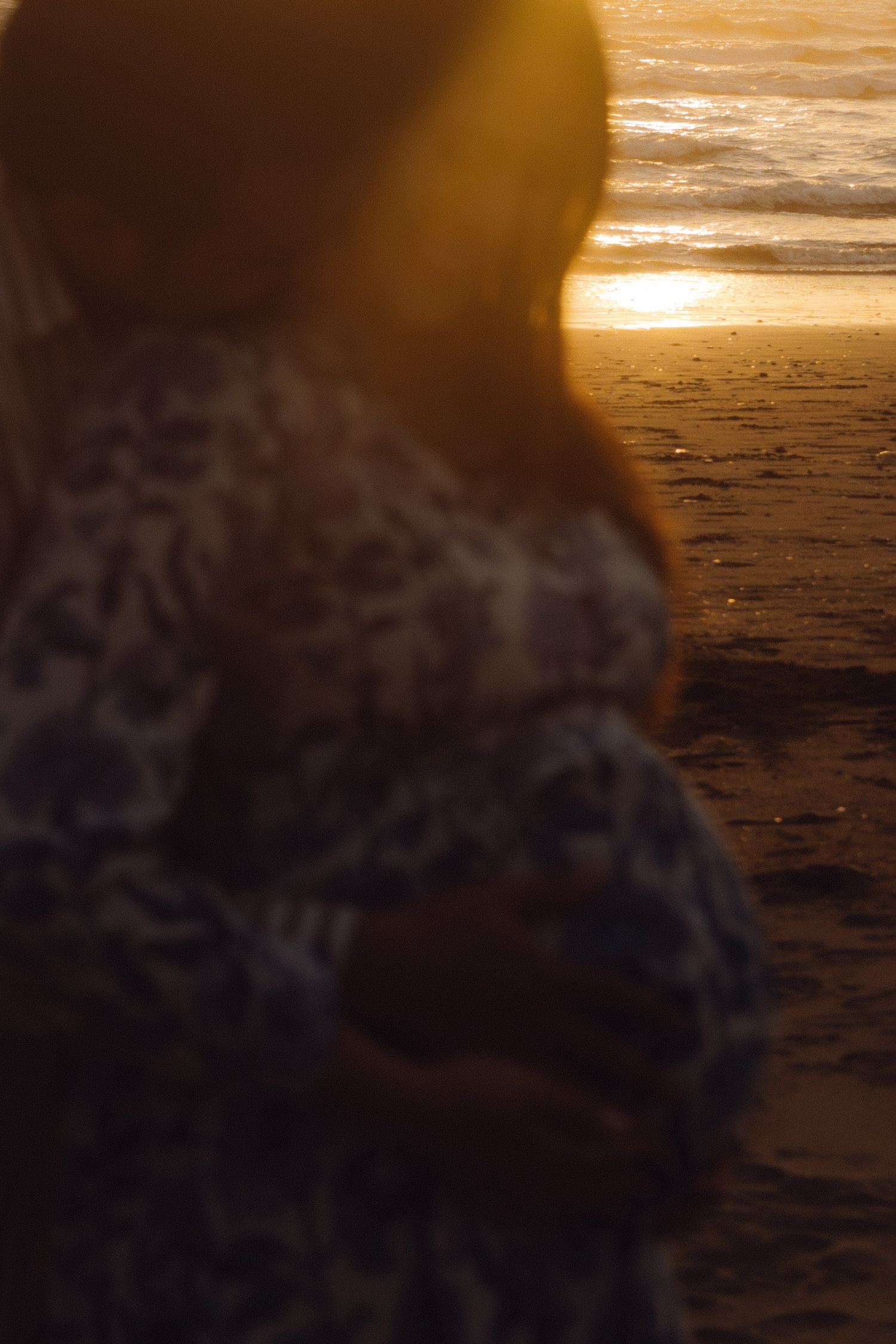 Blurred close-up of pregnant woman in floral dress standing by ocean shore during warm golden sunset.
