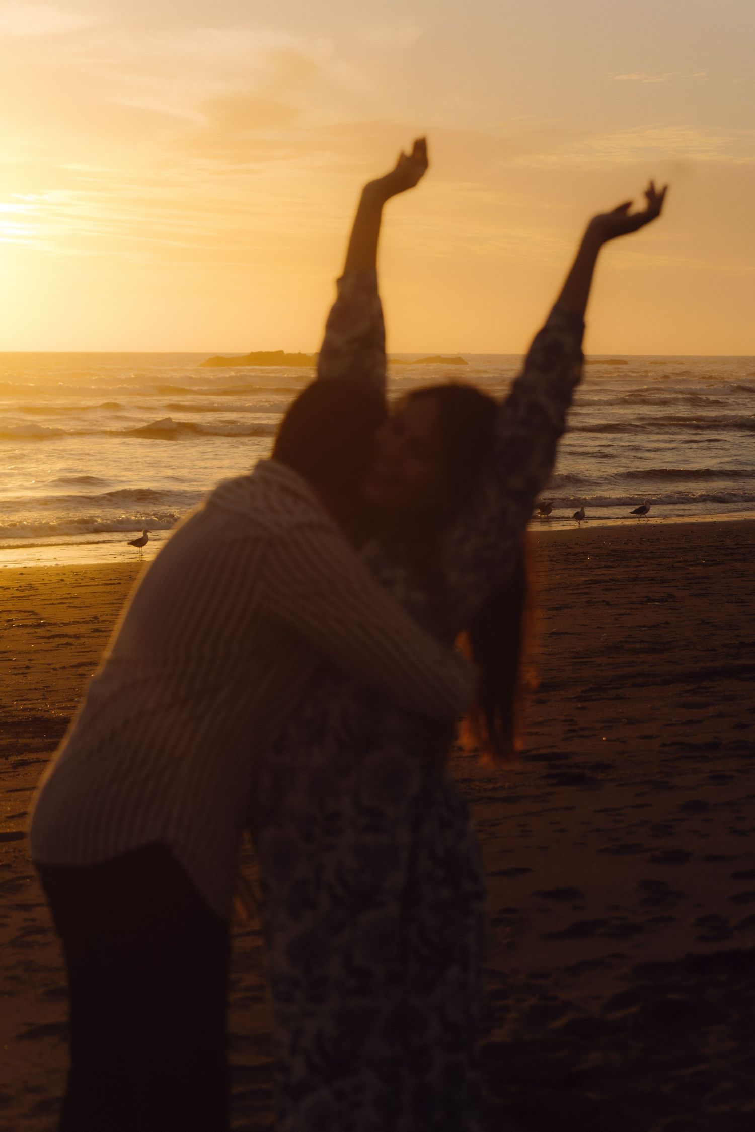 Silhouetted pregnant couple on beach at sunset, woman raising arms joyfully while partner embraces her.