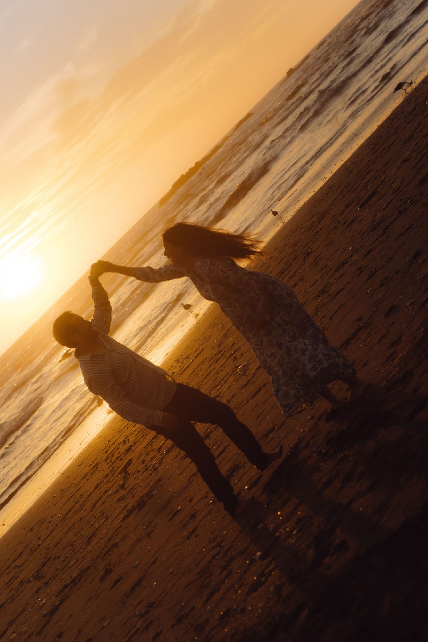 Silhouette of expecting couple holding hands on wet sandy beach during vibrant golden ocean sunset.