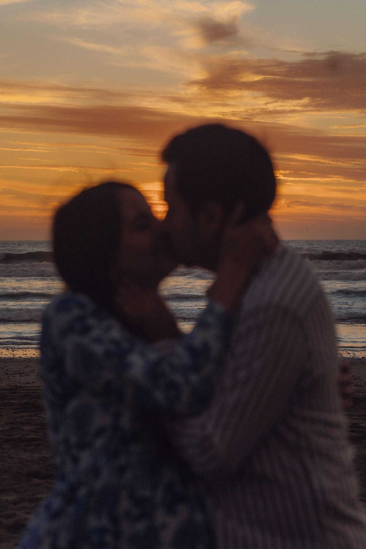 Expecting couple sharing a romantic kiss on the beach against a dramatic colorful sunset sky.