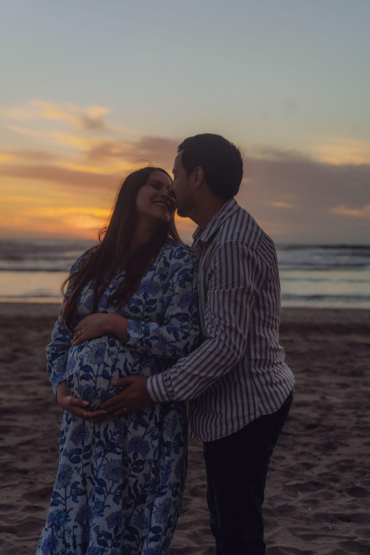 Man kissing pregnant partner on cheek while cradling her baby bump on beach at sunset.