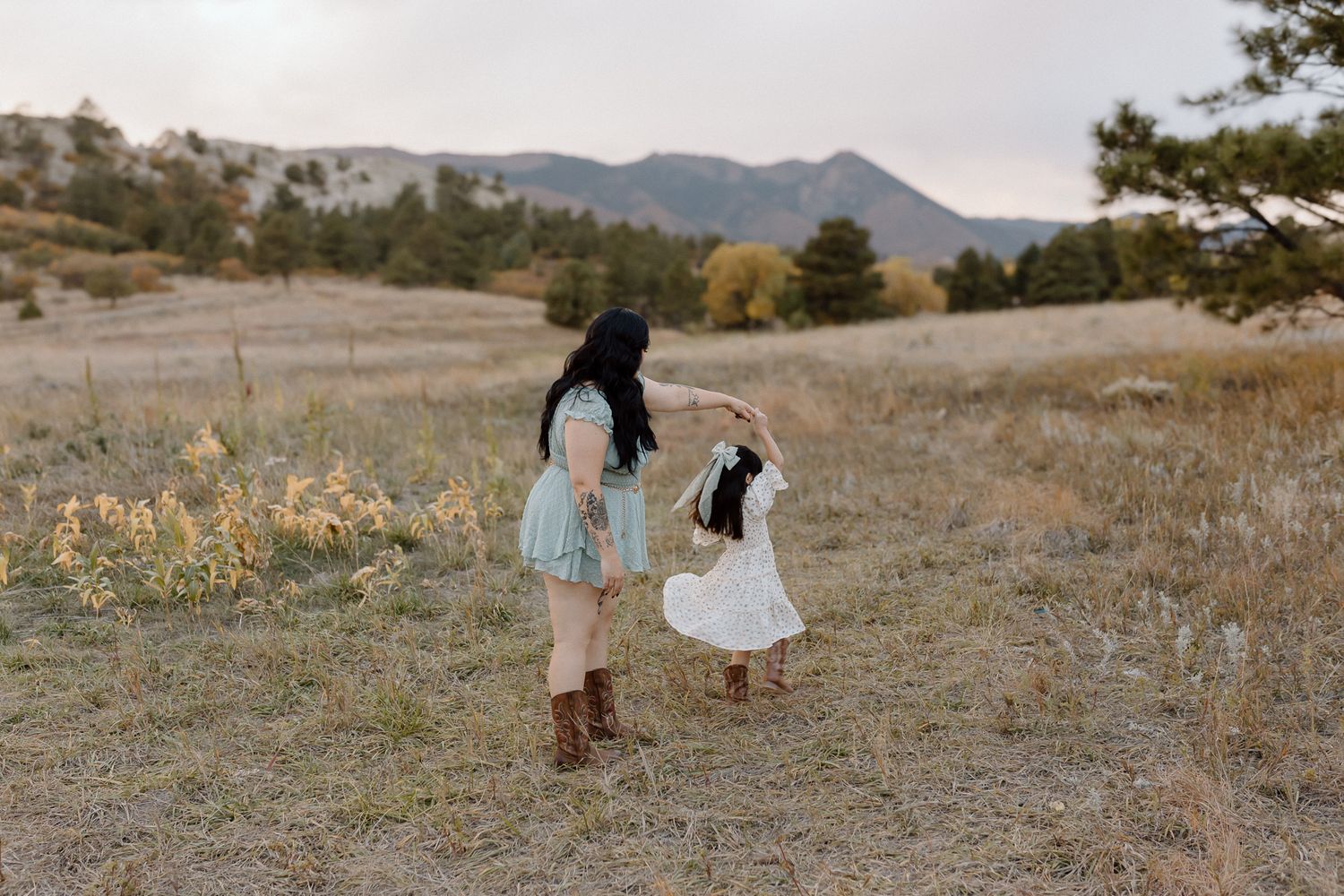 Two people in flowy dresses stand together in a mountain meadow at dusk with mountains in the distance.
