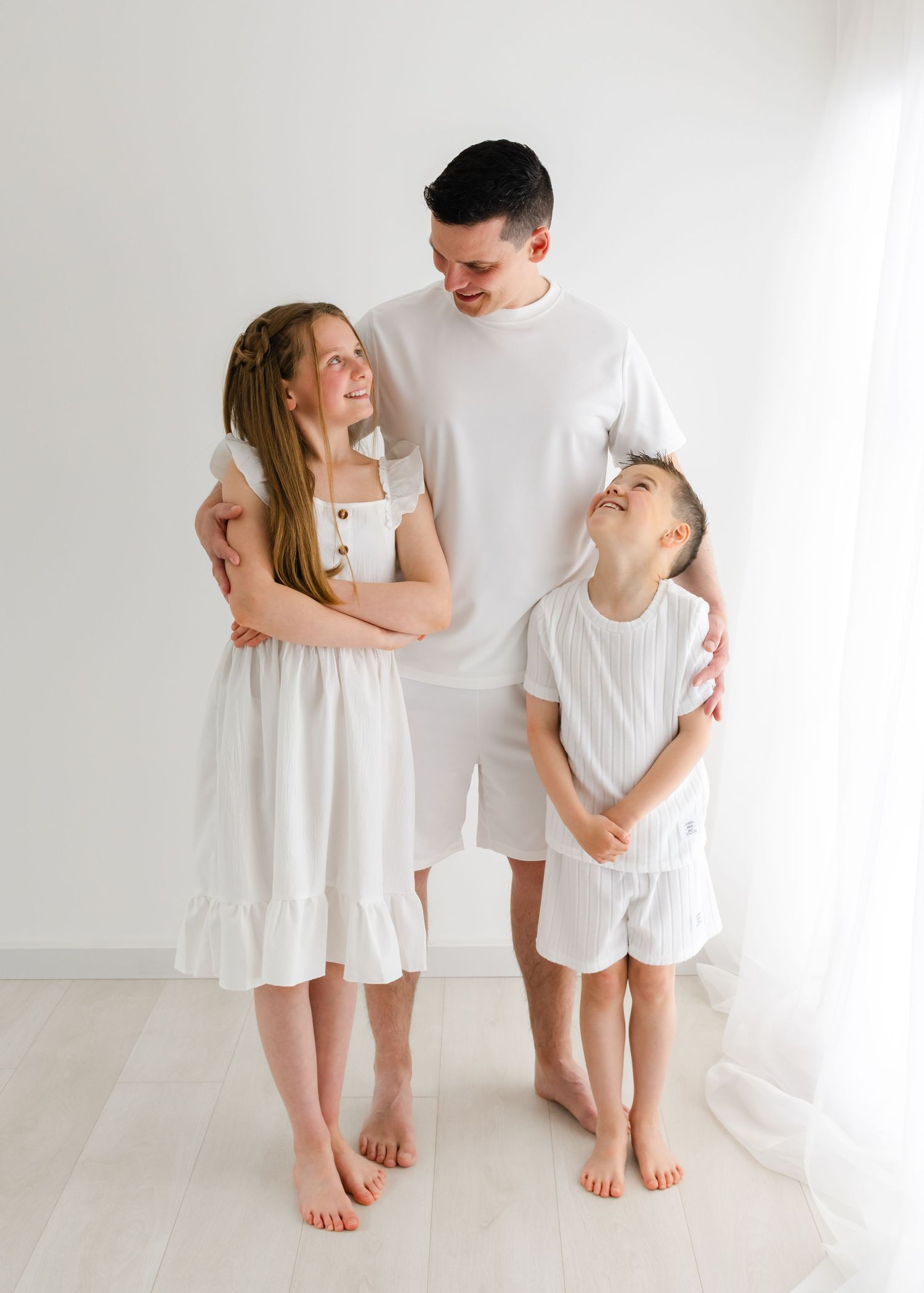Three people wearing matching white outfits stand together barefoot against a minimalist white wall in a studio setting.