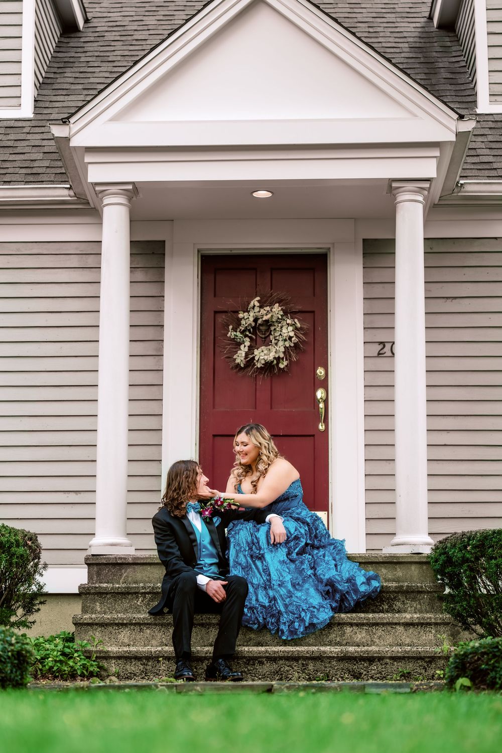 Prom couple sitting on front steps, woman in blue gown, man in tuxedo, red door backdrop.