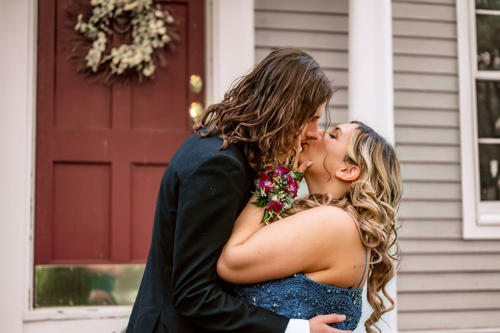 Couple sharing a romantic kiss in formal attire outside a house with a red door and wreath.