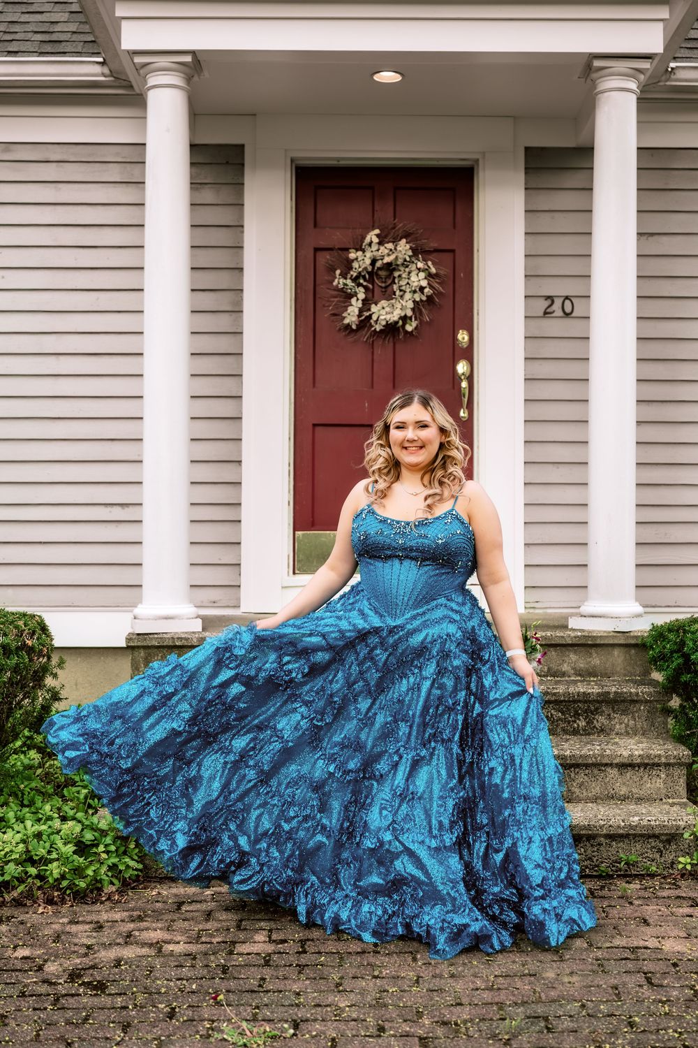 Young woman in a flowing teal ball gown poses on brick steps before a classic home with red door.