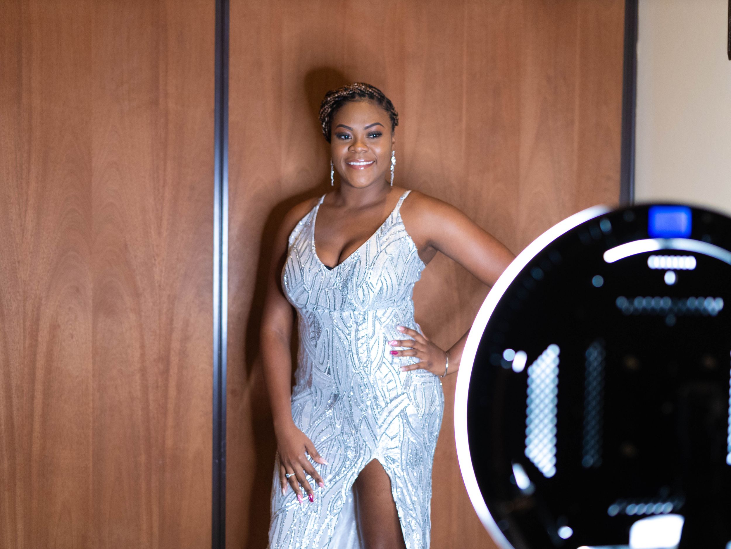 A person poses in a sparkly white sequined dress with a side slit against a wooden wall near a ring light.