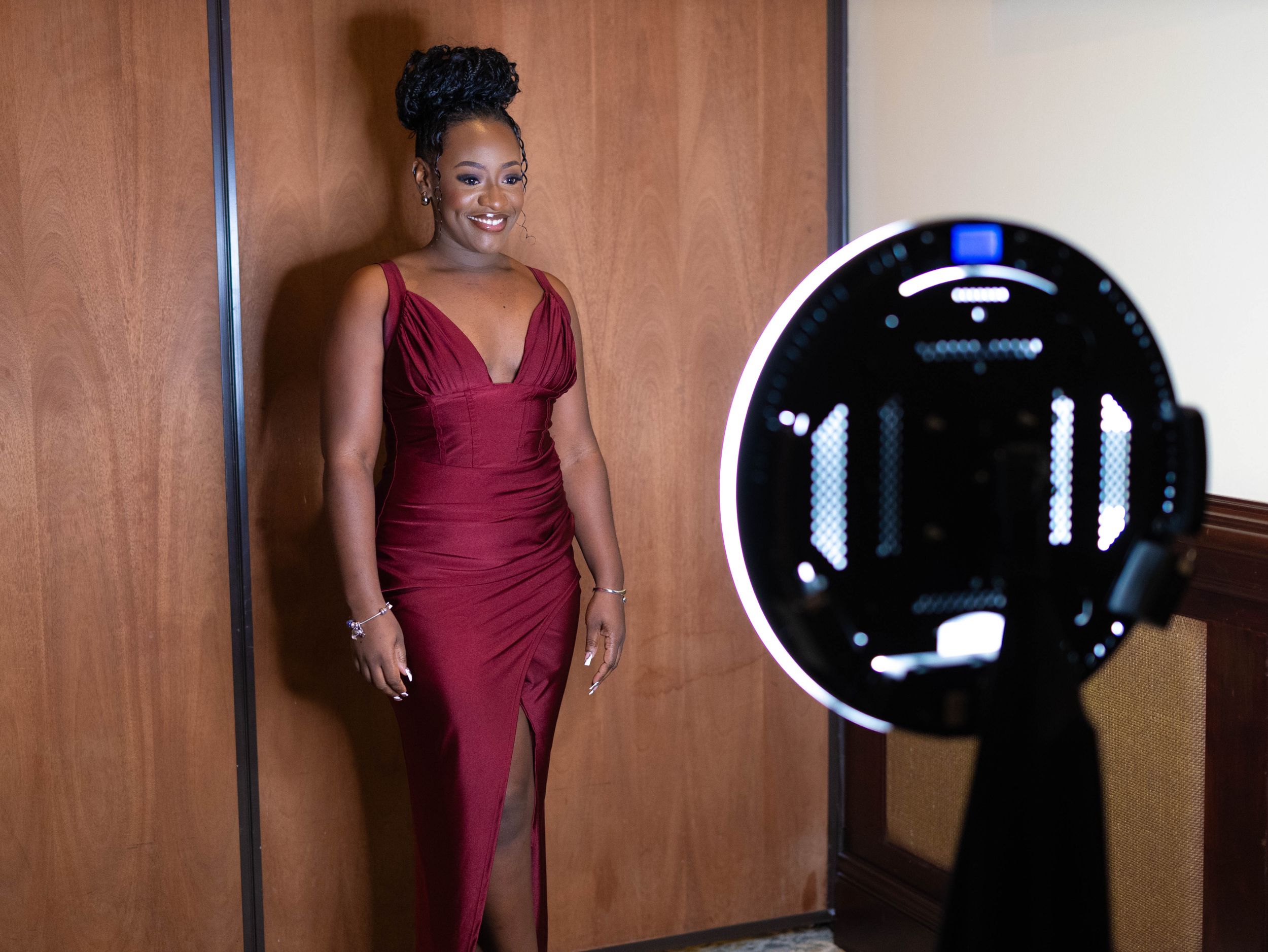 Behind the scenes photo showing a maroon evening gown with lighting equipment and wooden wall backdrop.