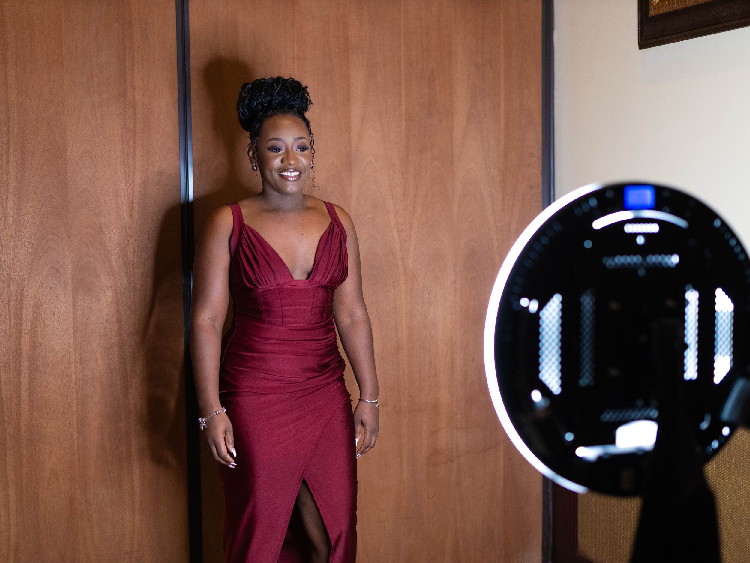 A woman in a burgundy formal dress poses next to a ring light in a studio setting.