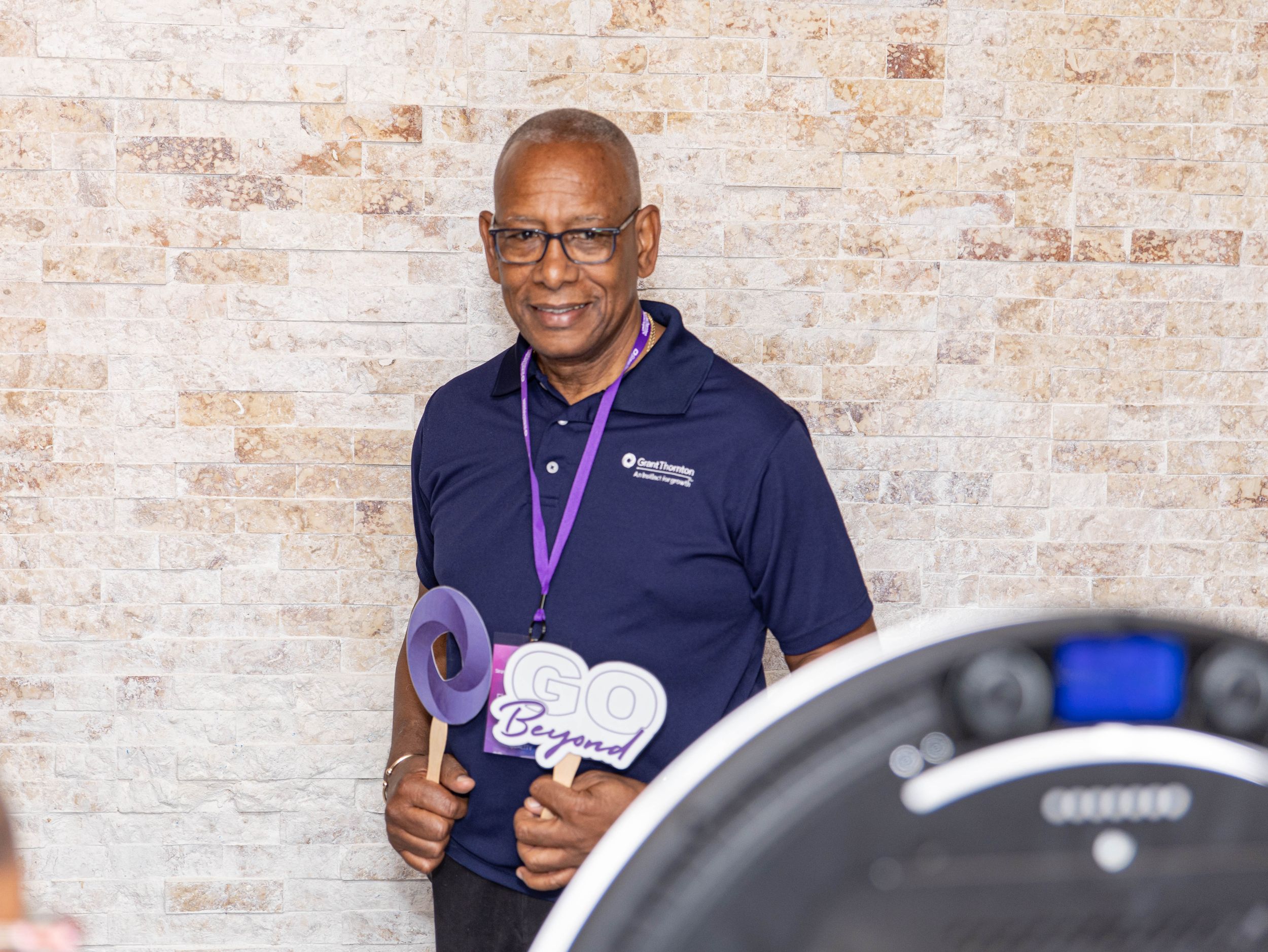 Someone wearing glasses and a navy blue pullover gives a thumbs up while standing near fitness equipment.