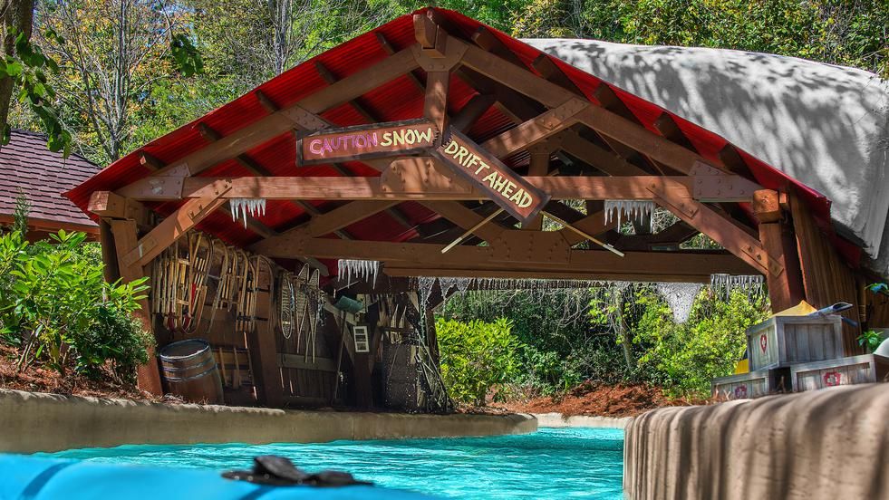 A rustic wooden footbridge with red trim spans over bright turquoise water at a theme park attraction.