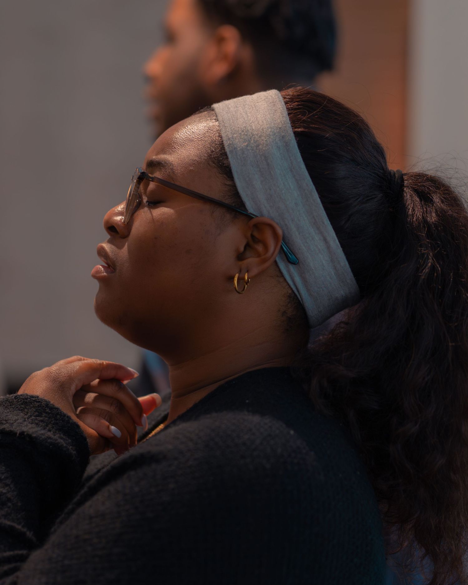 A person in gray attire standing with hands clasped in prayer beneath decorative lighting.