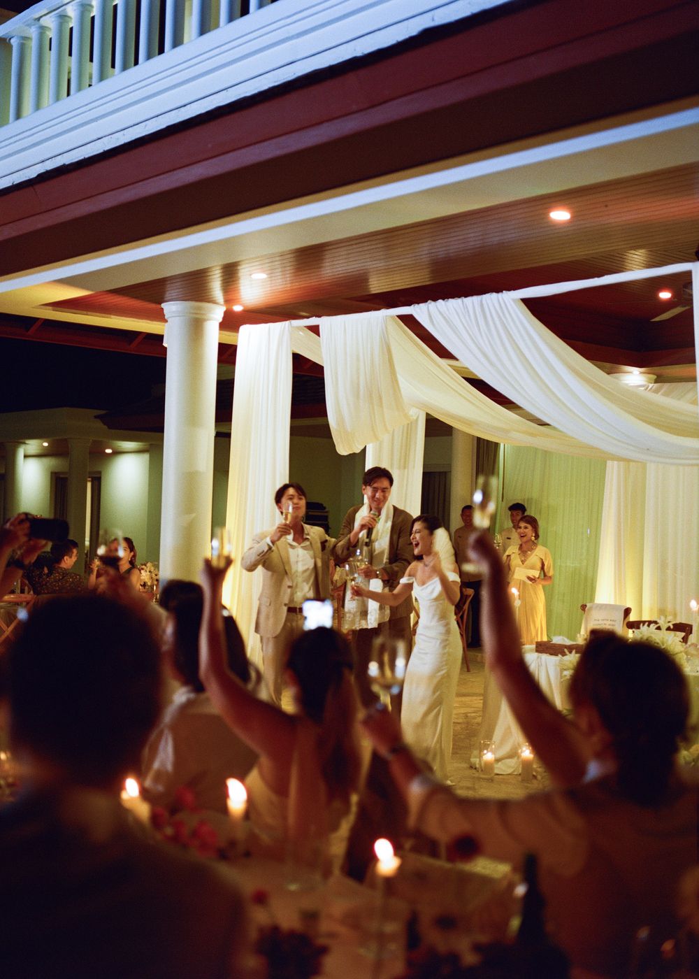 Wedding reception toast with bride and groom under elegant white draping, guests raising glasses in candlelit outdoor venue.