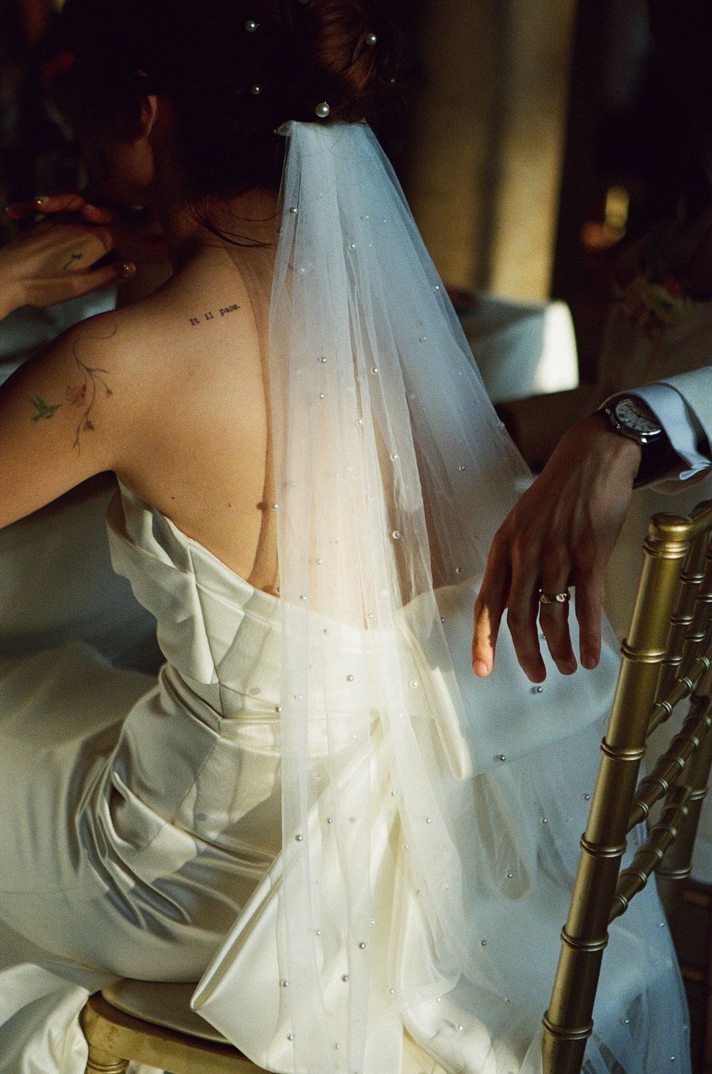 Bride in white satin gown with pearl-adorned veil and back tattoos, seated on gold chiavari chair at wedding.