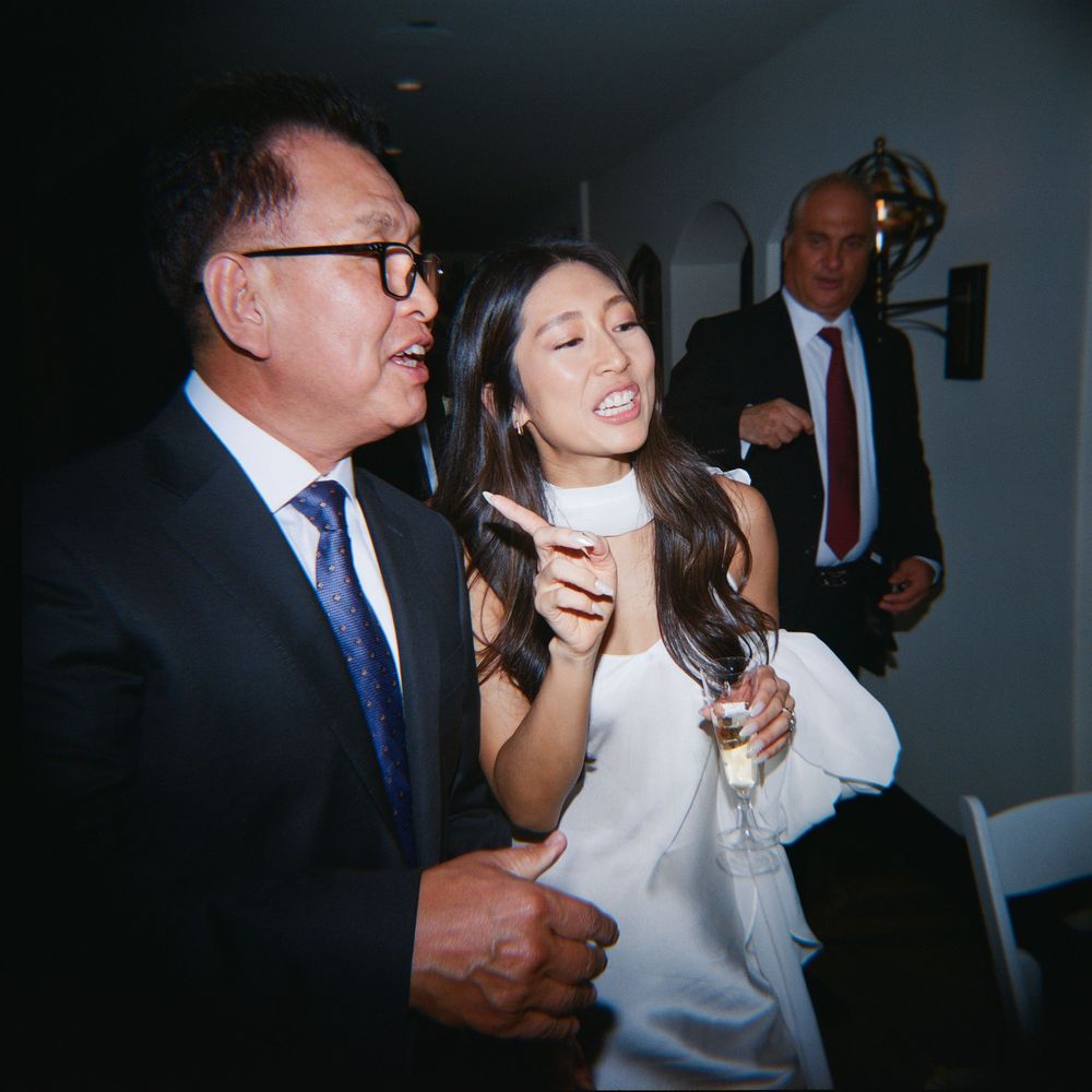 Two guests in formal attire chat at an elegant indoor event, woman in white holds champagne glass.