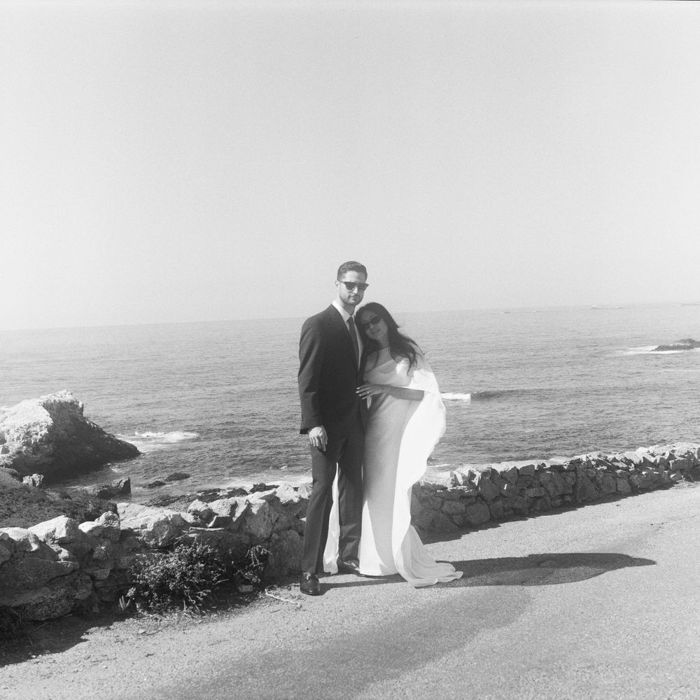 Black and white photo of a couple in formal wear posing by a rocky ocean coastline on a sunny day.
