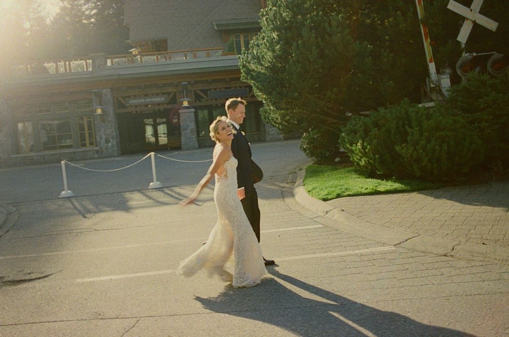 Bride and groom in wedding attire crossing a sunlit street near a railroad crossing sign.
