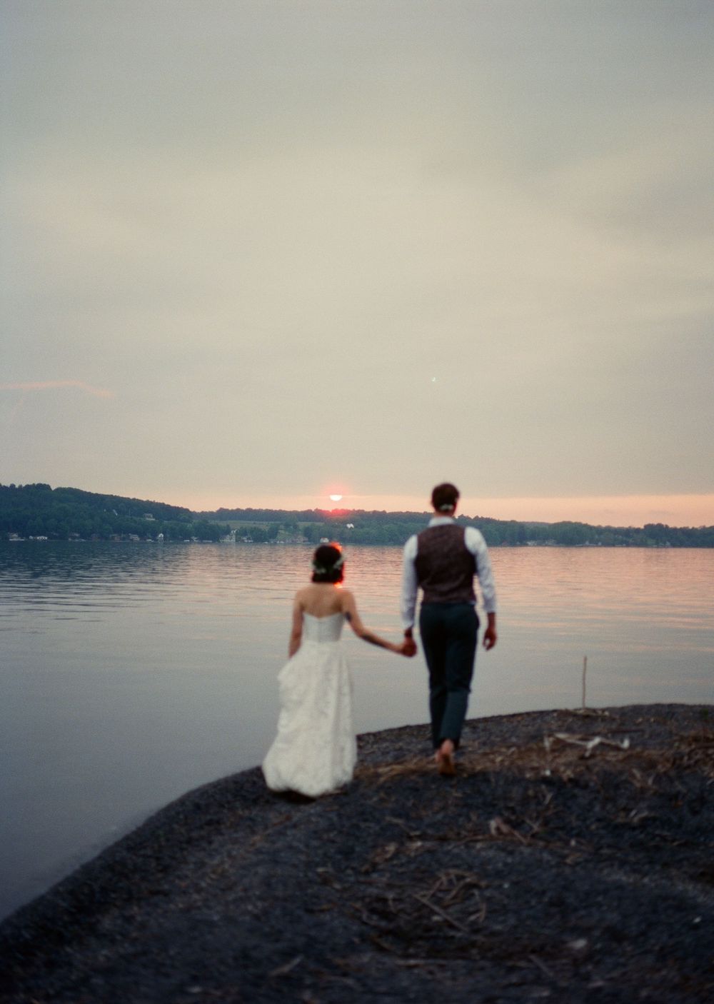 Couple stands silhouetted against pink sunset sky on rocky shoreline of lakeside beach.