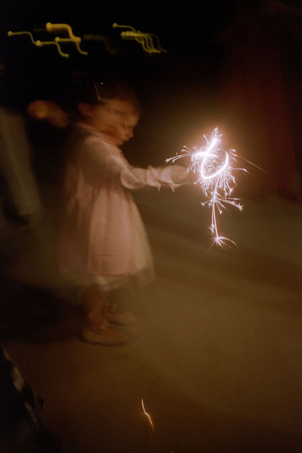 A small child in a white dress holds a glowing sparkler at night creating a magical moment.