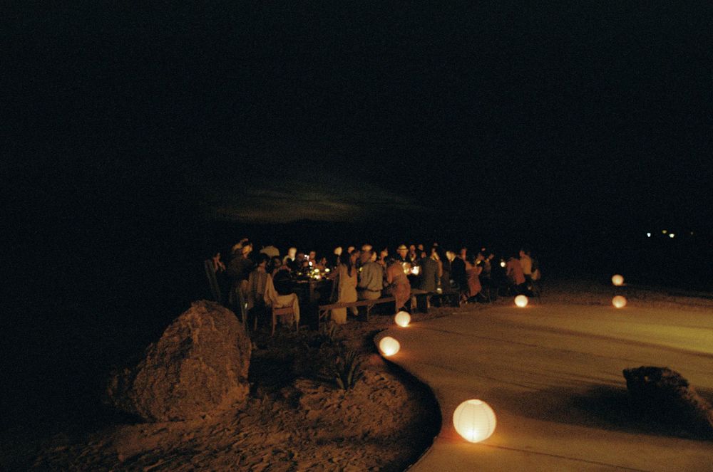 Outdoor nighttime gathering with glowing round lanterns lining a sandy path, guests mingling under a dark sky.