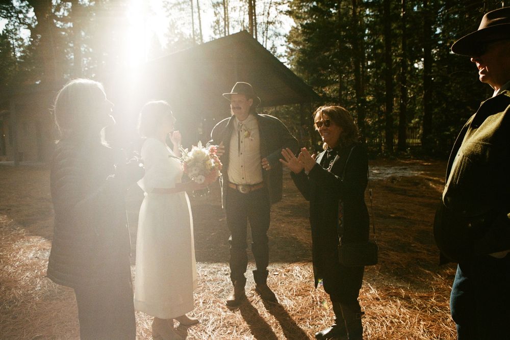 Outdoor wedding ceremony in sunlit forest with bride, groom, and guests gathered near a tent.