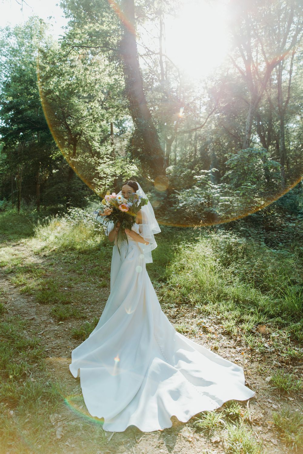 A figure in a flowing white wedding gown holds a bouquet in a sunlit forest setting.