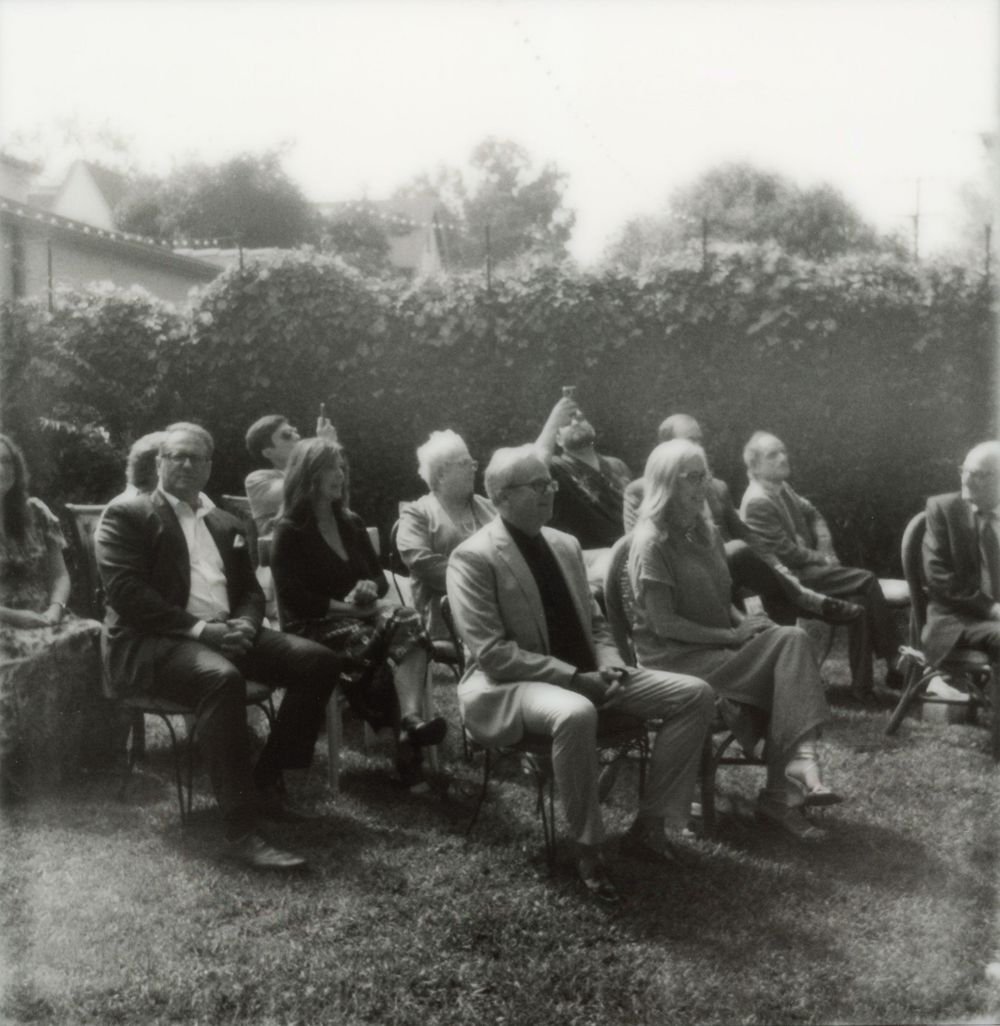 A vintage black and white photo shows guests seated in rows at an outdoor gathering.