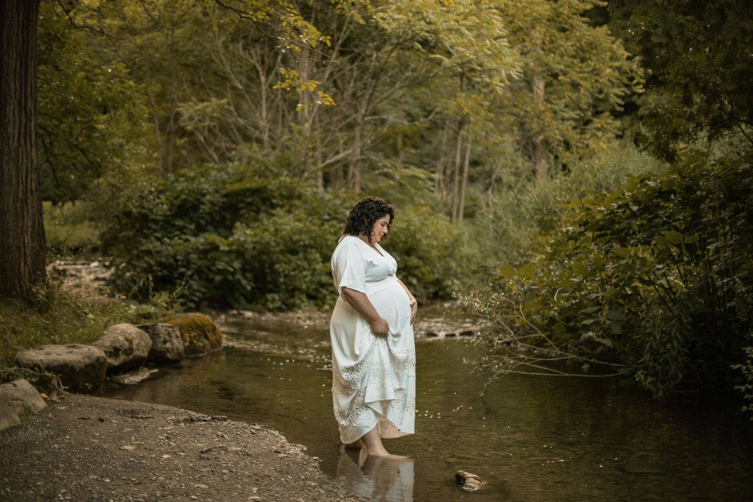 A pregnant woman in a black blazer basking in sunlight against a blue sky background, touching her belly with gentle serenity.