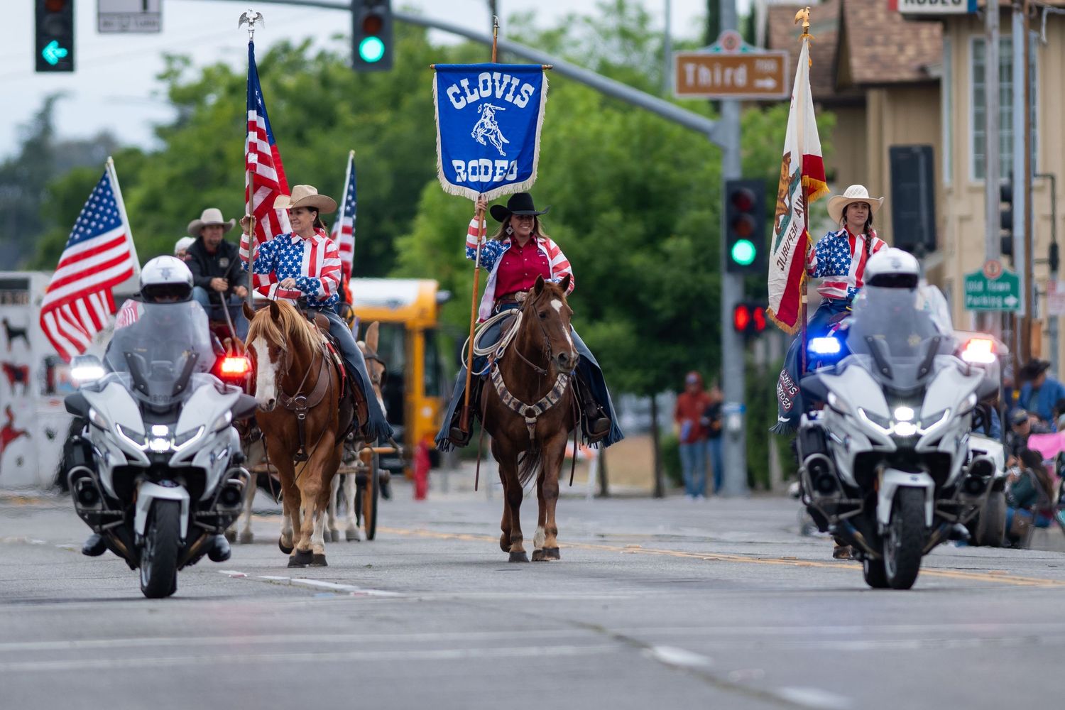 Parade procession with motorcycles and mounted riders on a city street with American flags displayed.
