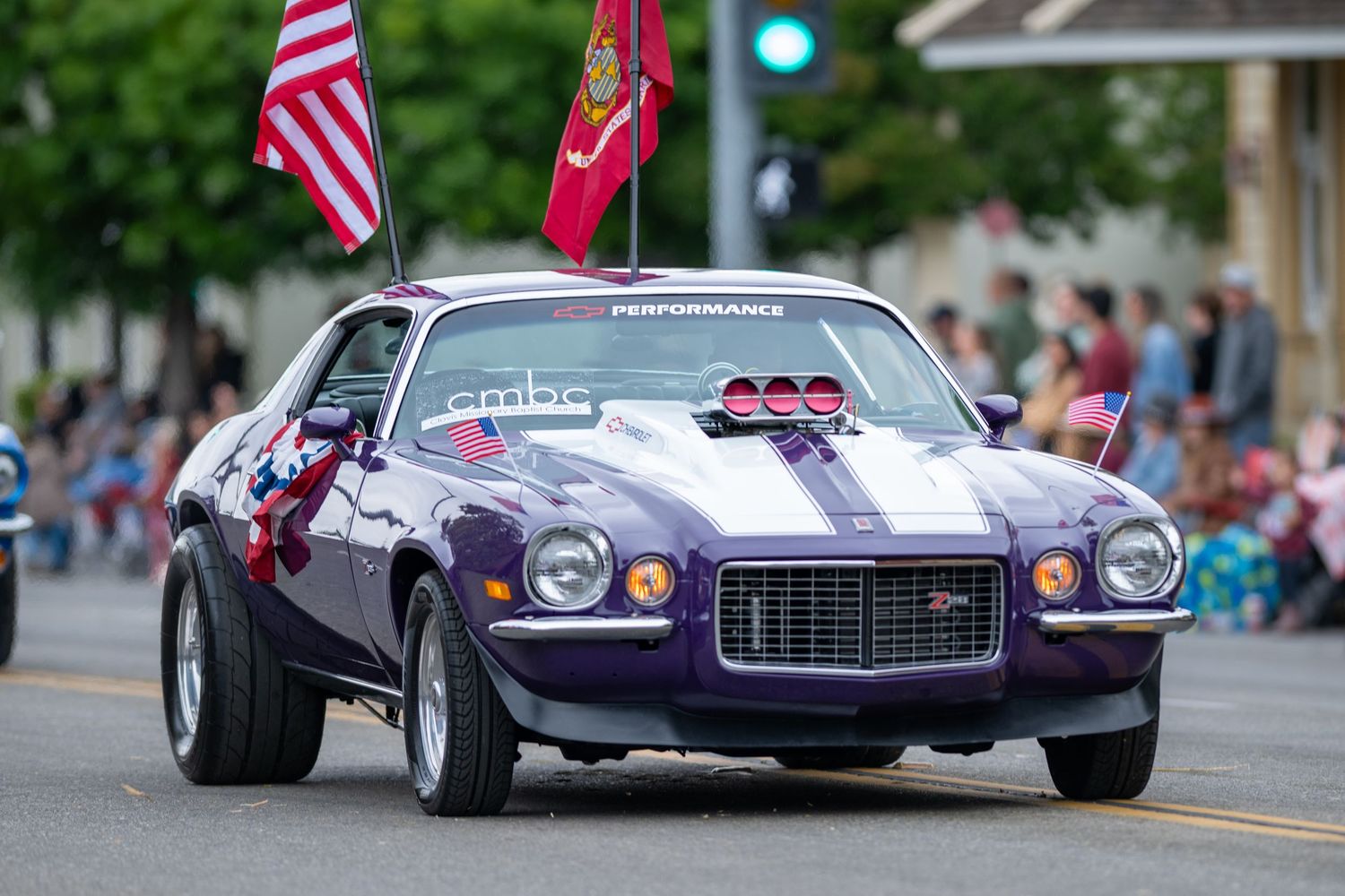 Purple Chevrolet Camaro muscle car with American flags displayed during a parade.