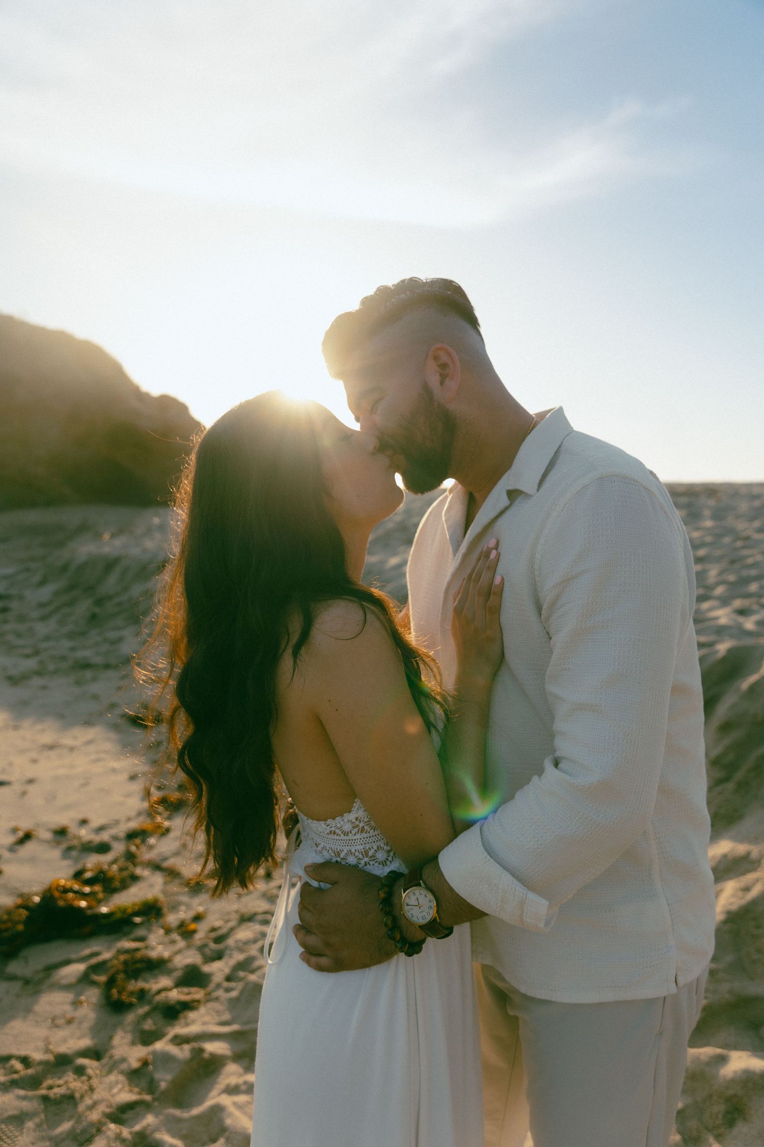 A romantic beach kiss at sunset with mountains and golden sunlight shining between a couple in white wedding attire.