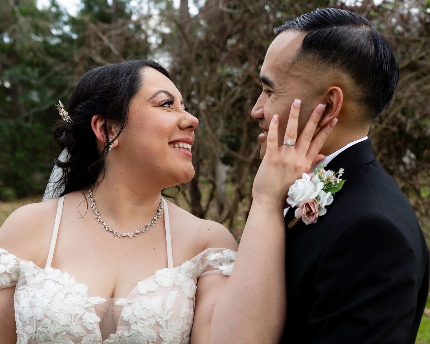 A newlywed couple enjoys a candid moment outdoors as they pose together on grassy lawn near wooded area.