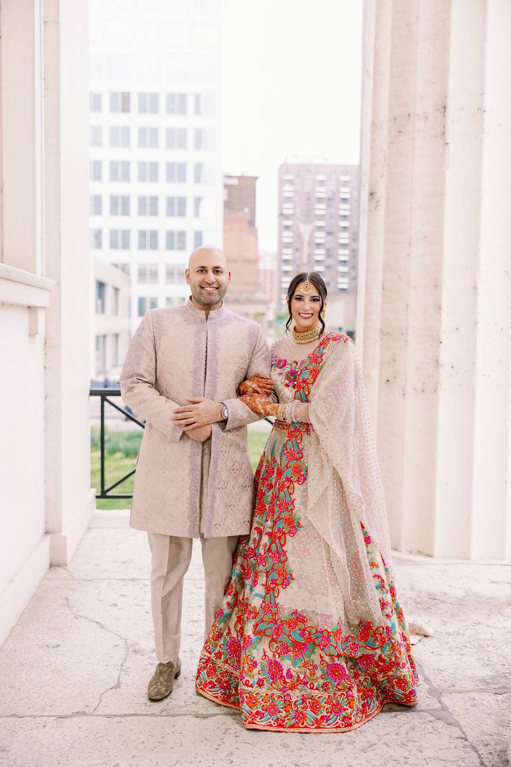 A couple in South Asian wedding attire stands between marble columns with a cityscape backdrop.